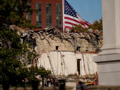 A US flag flies in the background as the ruins of the facade of the East Wing can be seen in the foreground